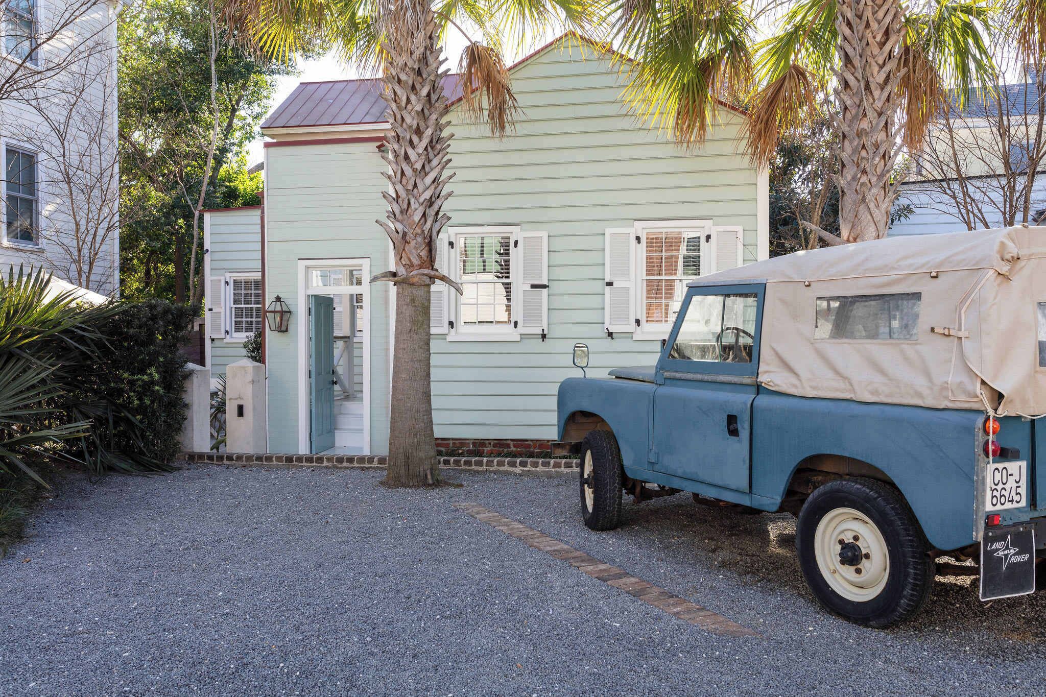 Un vieux Land Rover bleu est garé devant une maison vert clair aux volets blancs.
