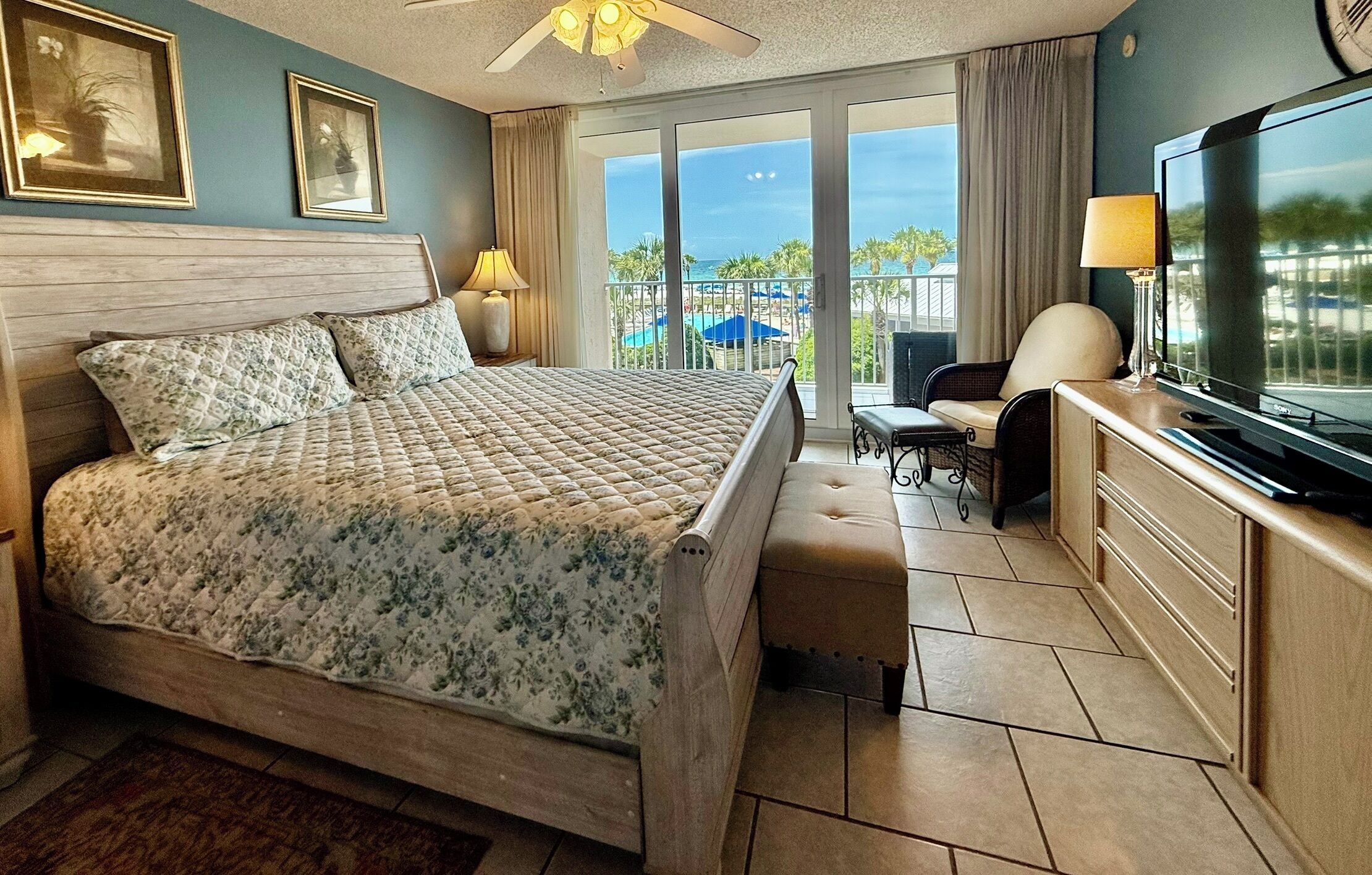 A hotel room with a bed, a television, a desk with a chair, and a balcony with a view of the ocean.