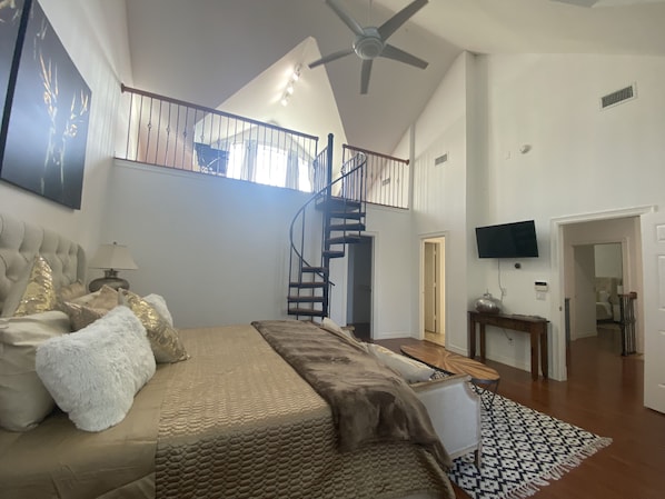 Um quarto espaçoso com uma cama grande, um ventilador de teto, uma escada e uma televisão fixada na parede.