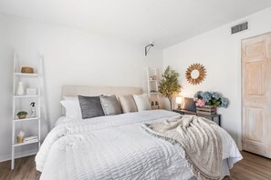 Un dormitorio con cama, mesita de noche, lámpara, estante con objetos decorativos y una puerta.