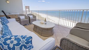 En balkon med en hvid sofa, fletstole og et bord med en flaske og glas. Der kan ses en strandudsigt gennem gelænderet.