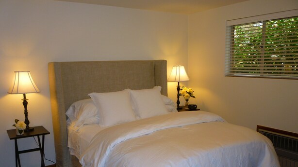 A neatly made bed with white linens, two bedside lamps, and a window with blinds.