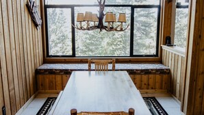 Interior de cabana de madeira com uma grande mesa de jantar, bancos, um candelabro e uma janela com vista para árvores.