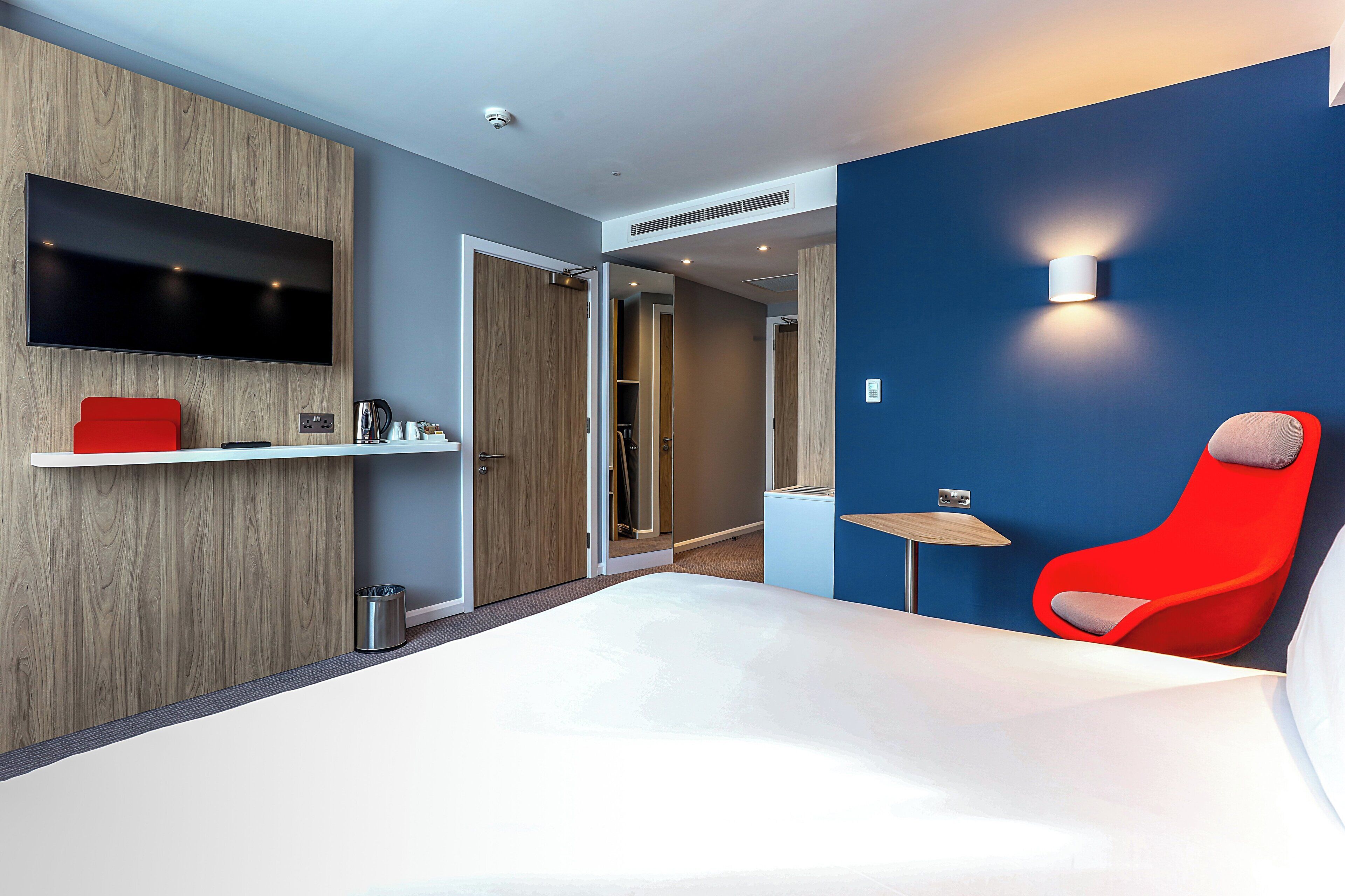 A modern hotel room with a large bed, a red chair, a TV mounted on the wall, and a small table with a lamp.