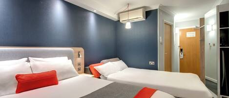 Une chambre d’hôtel avec deux lits, un oreiller rouge, une couverture grise et une tête de lit blanche.