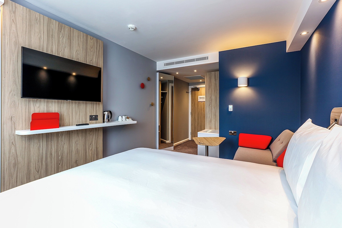 A modern hotel room with a large bed, a mounted TV, a small table, and a red accent chair.