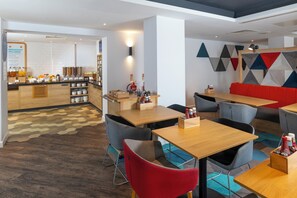 A modern dining area with wooden tables and chairs, a breakfast bar with various food items, and a geometric wall design.