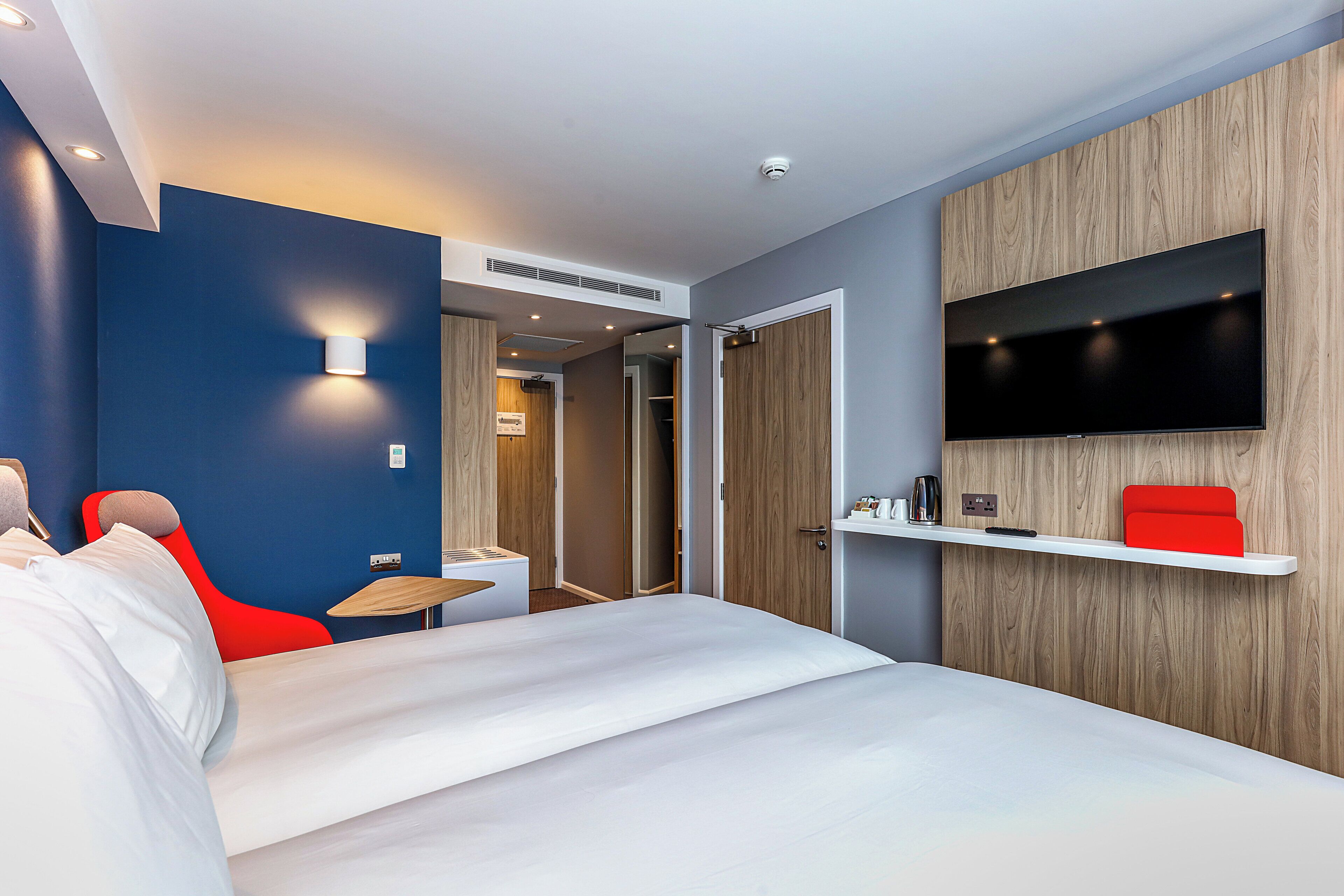 Een moderne hotelkamer met een bed, een rode stoel, een aan de muur bevestigde televisie en een klein tafeltje.