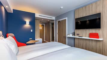 A modern hotel room with a bed, a red chair, a TV mounted on the wall, and a small table.