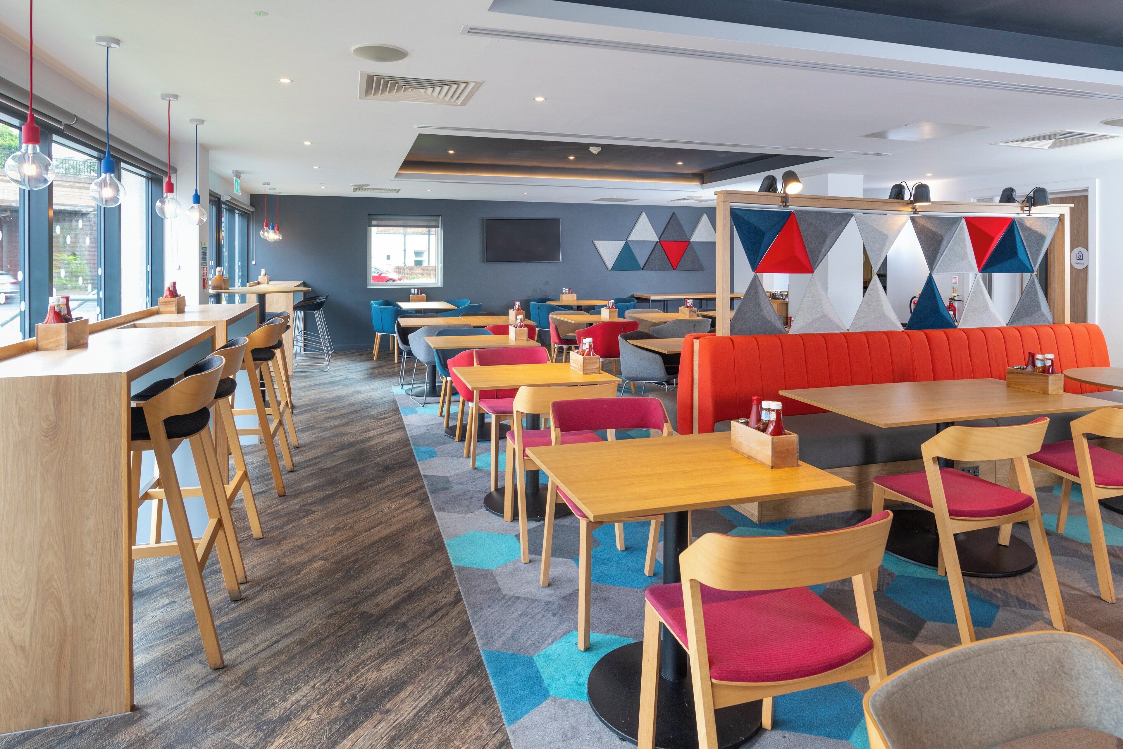 A modern restaurant interior with wooden tables and chairs, a colorful geometric wall decoration, and a large window providing natural light.