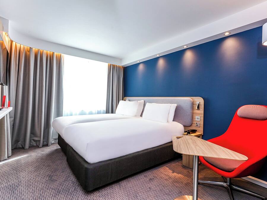 Una habitación de hotel con una cama grande, un escritorio, una silla, un televisor y un ventanal con cortinas.