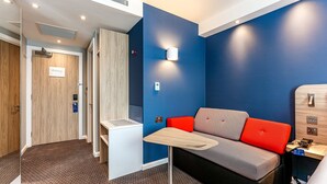A modern hotel room with a blue accent wall, a grey and red sofa, a small wooden table, and a bedside table with a lamp. - Holiday Inn Express Exeter - City Centre by IHG (Exeter)