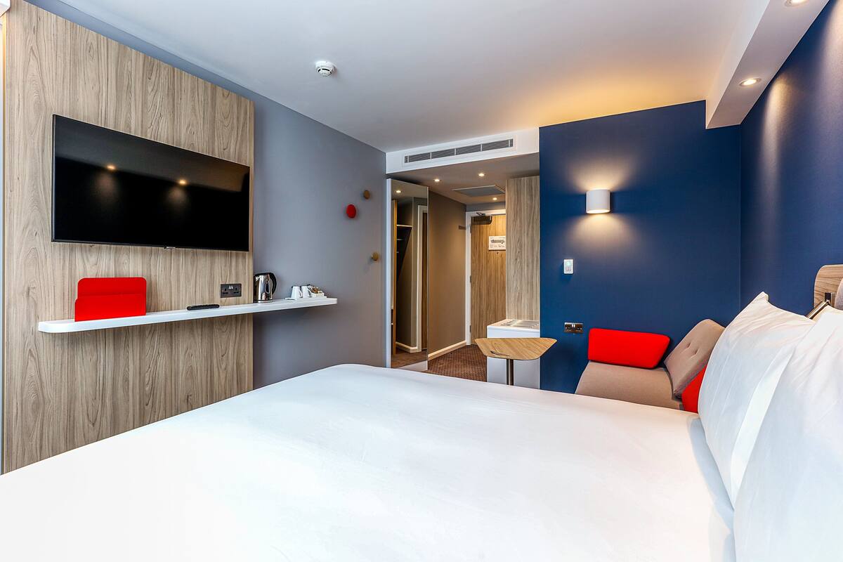 A modern hotel room with a large bed, a mounted TV, a small table, and a red accent chair.