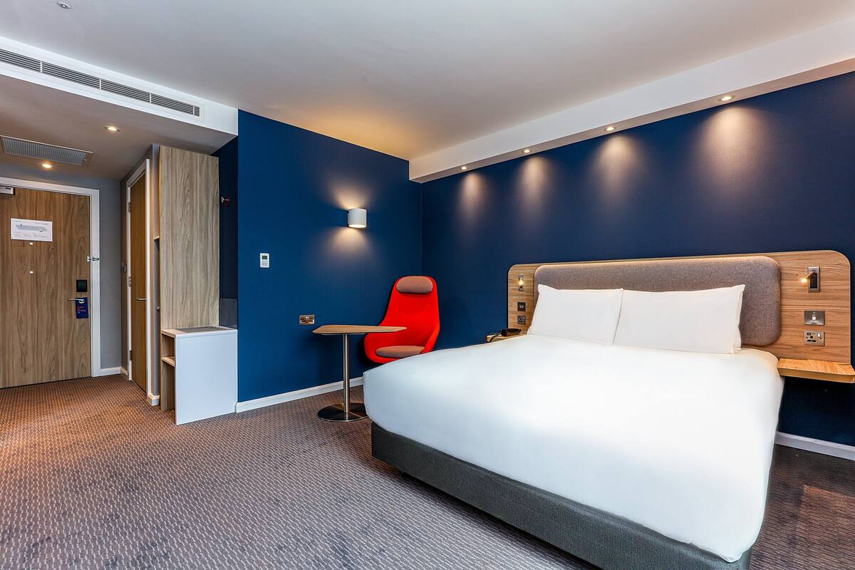 A modern hotel room with a large bed, a red chair, a small table, and a wooden wardrobe.