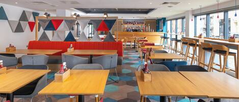 A modern restaurant with wooden tables and chairs, a bar area, and a colorful geometric wall design.