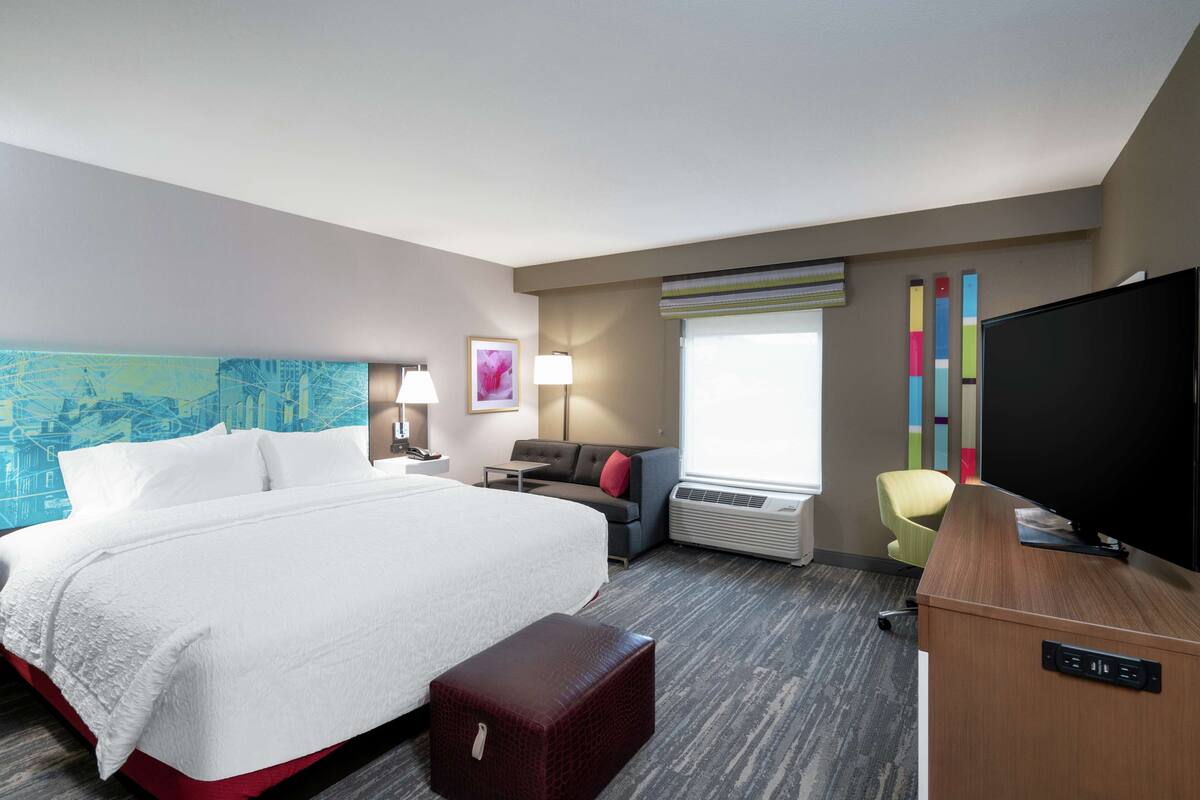 Une chambre d’hôtel avec un grand lit, un bureau avec un téléviseur à écran plat, un canapé, un fauteuil et une fenêtre avec des stores.