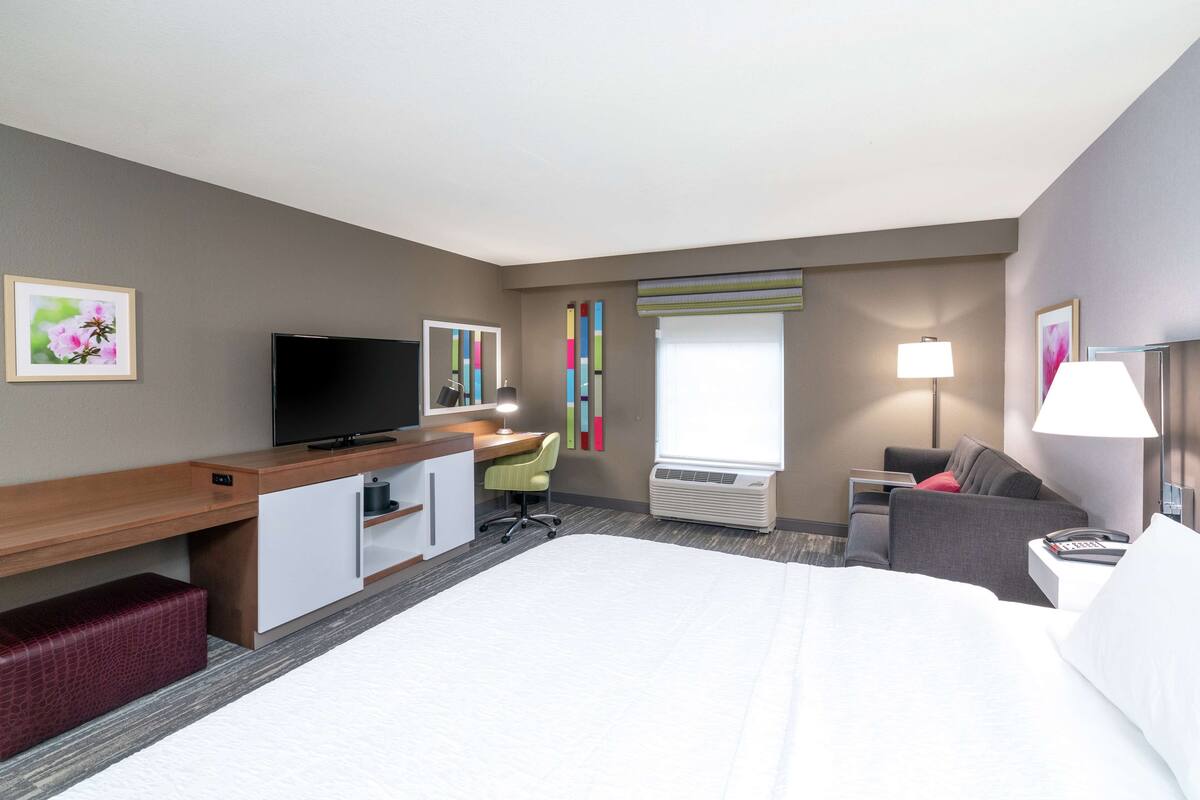 A hotel room with a large bed, a desk with a TV, a chair, a sofa, and a lamp.