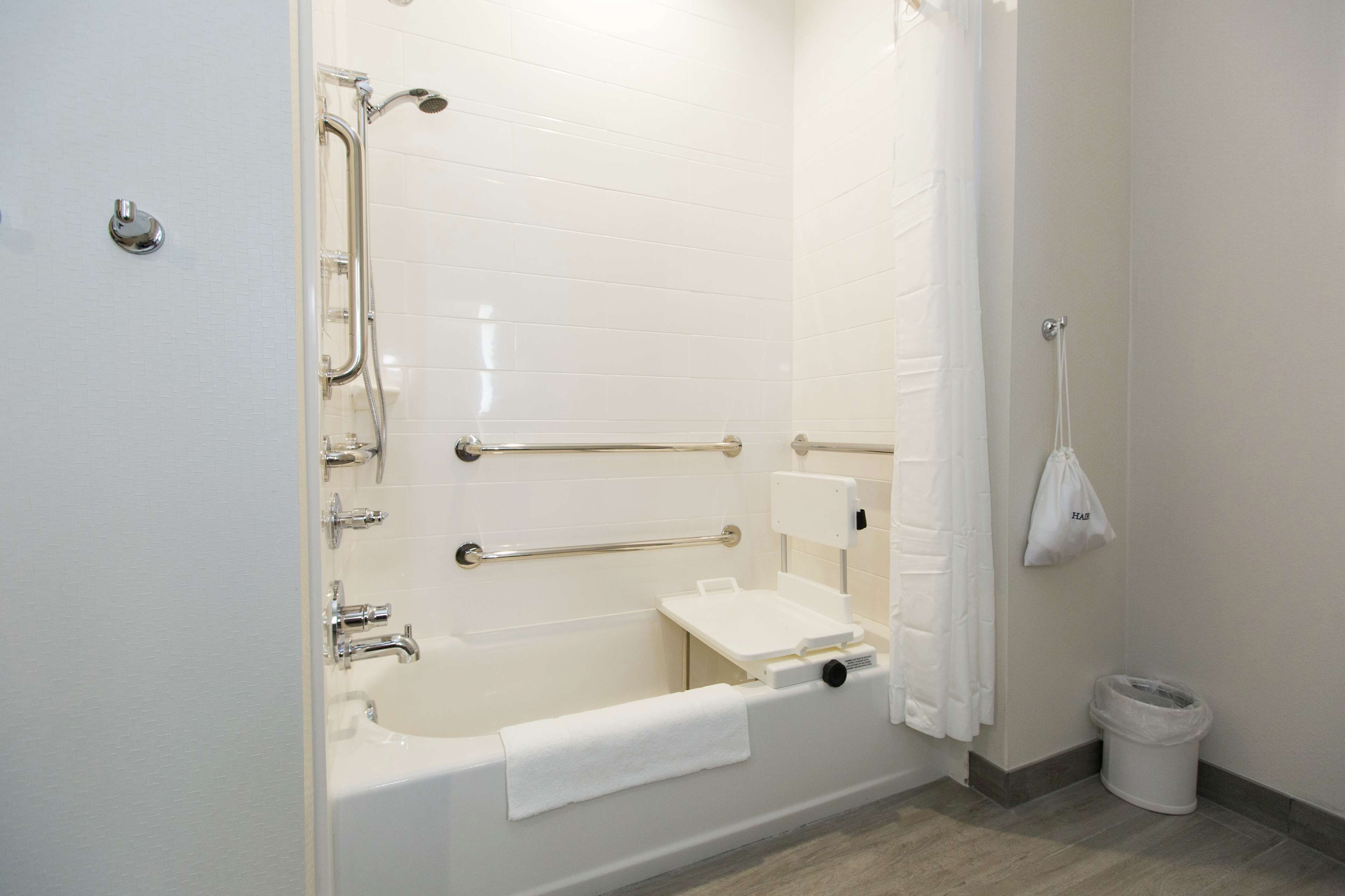 A modern bathroom with a bathtub, grab bars, a handheld showerhead, a towel rack, and a trash can.