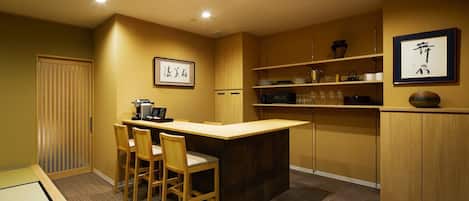 A compact kitchen with a wooden dining table and chairs, a coffee machine, and shelves with kitchenware.
