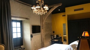 Ein Hotelzimmer mit einem großen Bett, einem Kronleuchter, einem Schreibtisch mit Stuhl, einem Fernseher und einem Fenster mit Vorhängen.