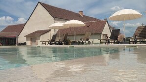 Ein Schwimmbecken mit Liegestühlen und Sonnenschirmen, ein Gebäude mit schrägem Dach und ein klarer Himmel.