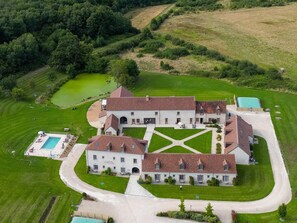 Un grand bâtiment à étages, doté d’une cour symétrique, entouré d’une végétation luxuriante et d’un petit étang.