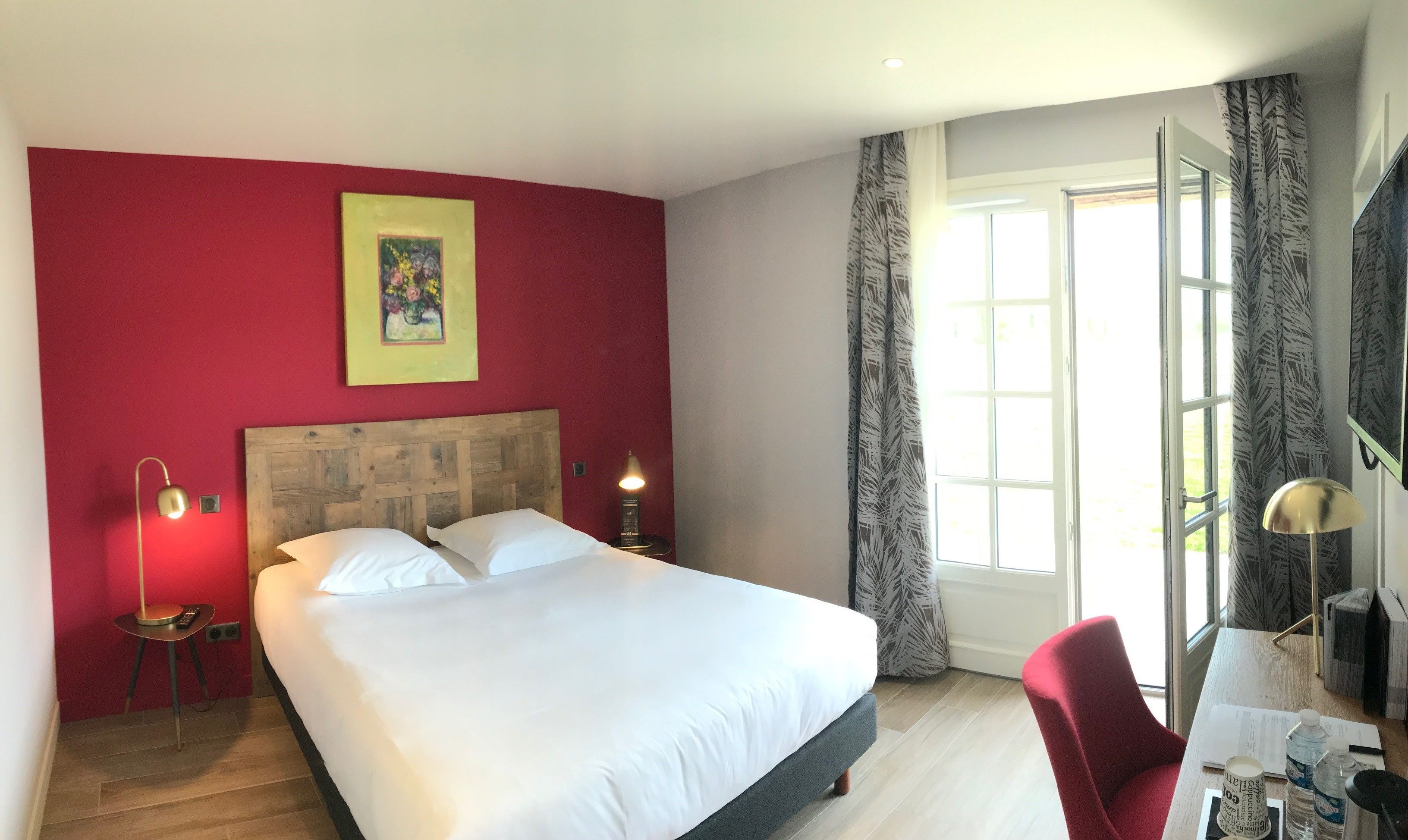 A hotel room with a large bed, a desk with a chair, a window with curtains, a painting on the wall, and a lamp.
