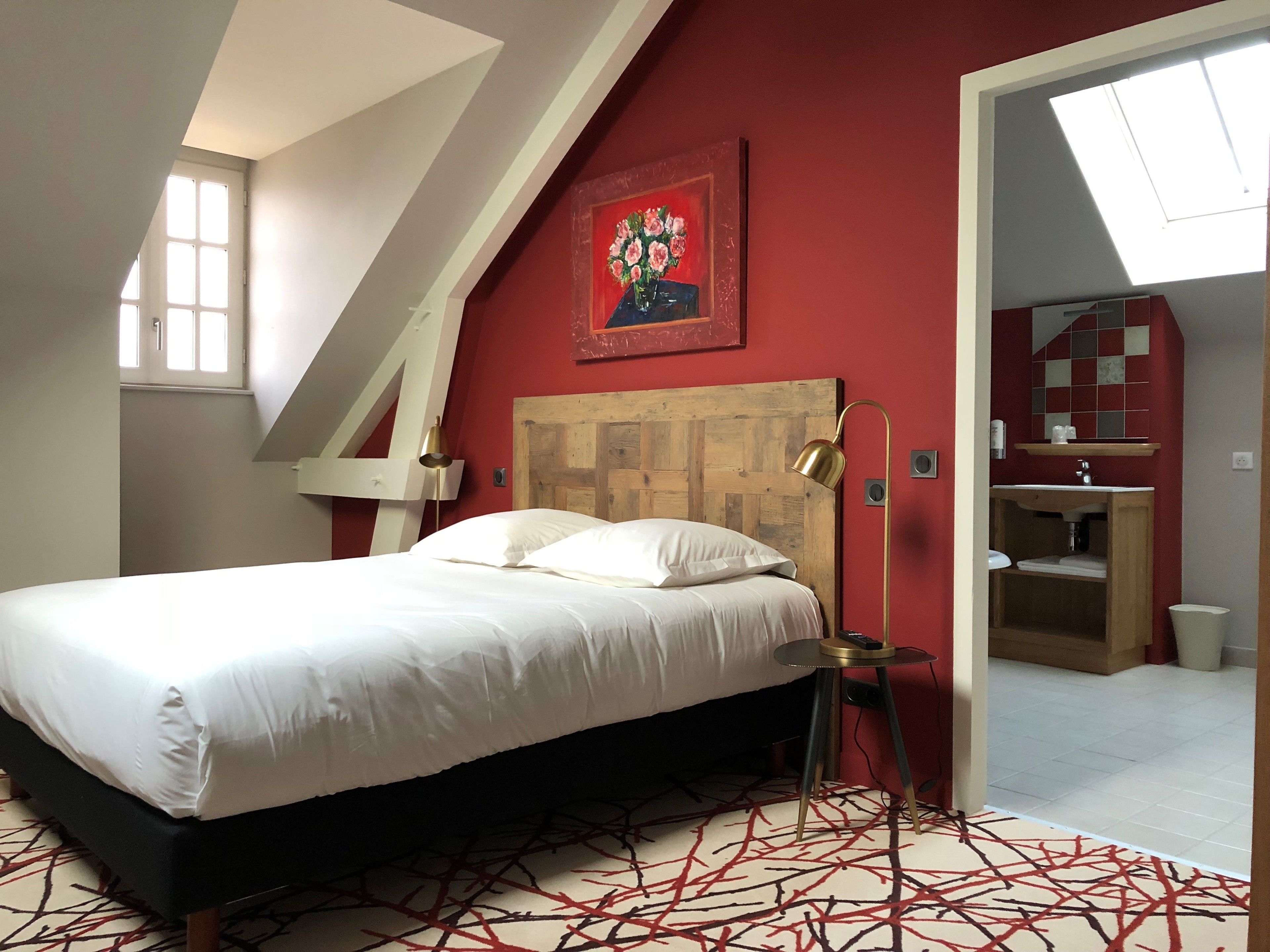 A bedroom with a bed, a wooden headboard, a painting, a bedside lamp, and a view of the bathroom.