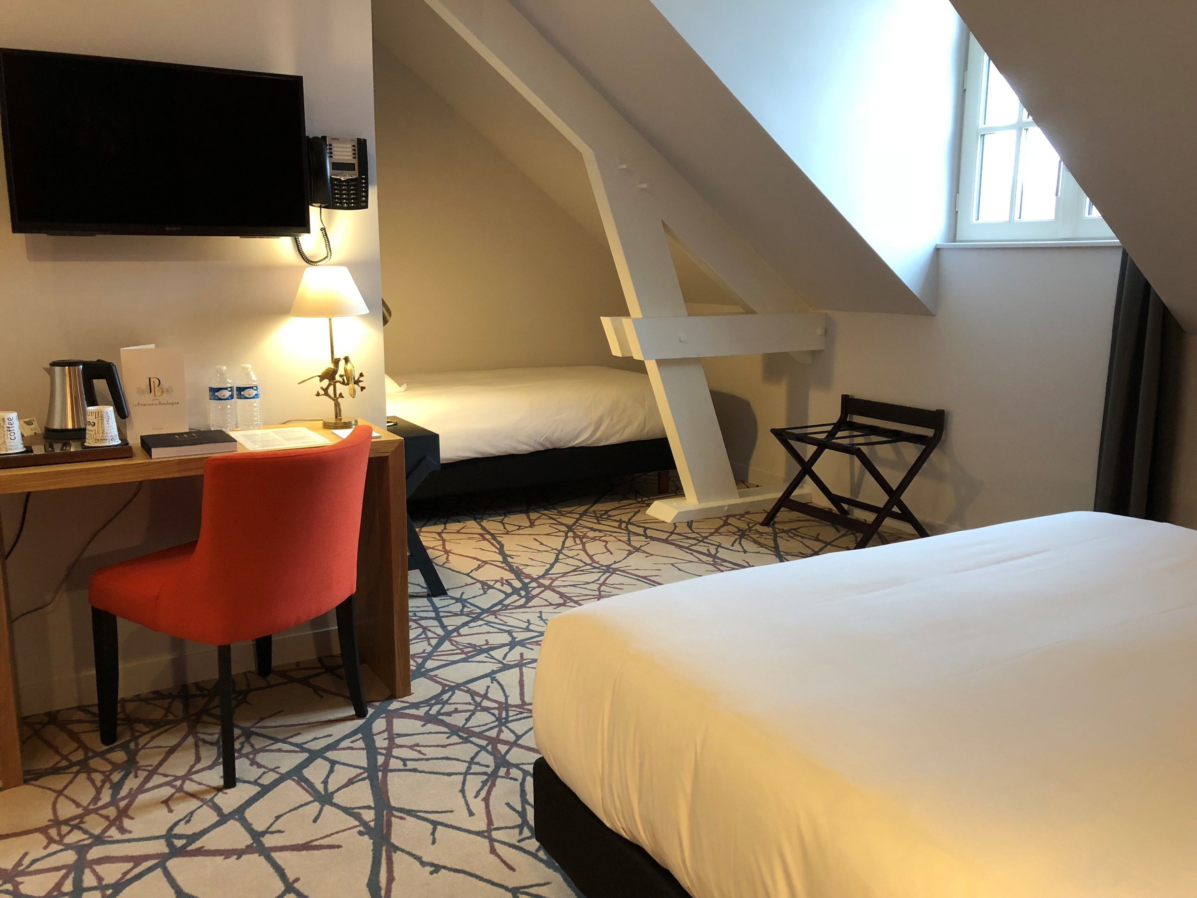 A hotel room with a desk, a red chair, a bed, a small table, and a window.