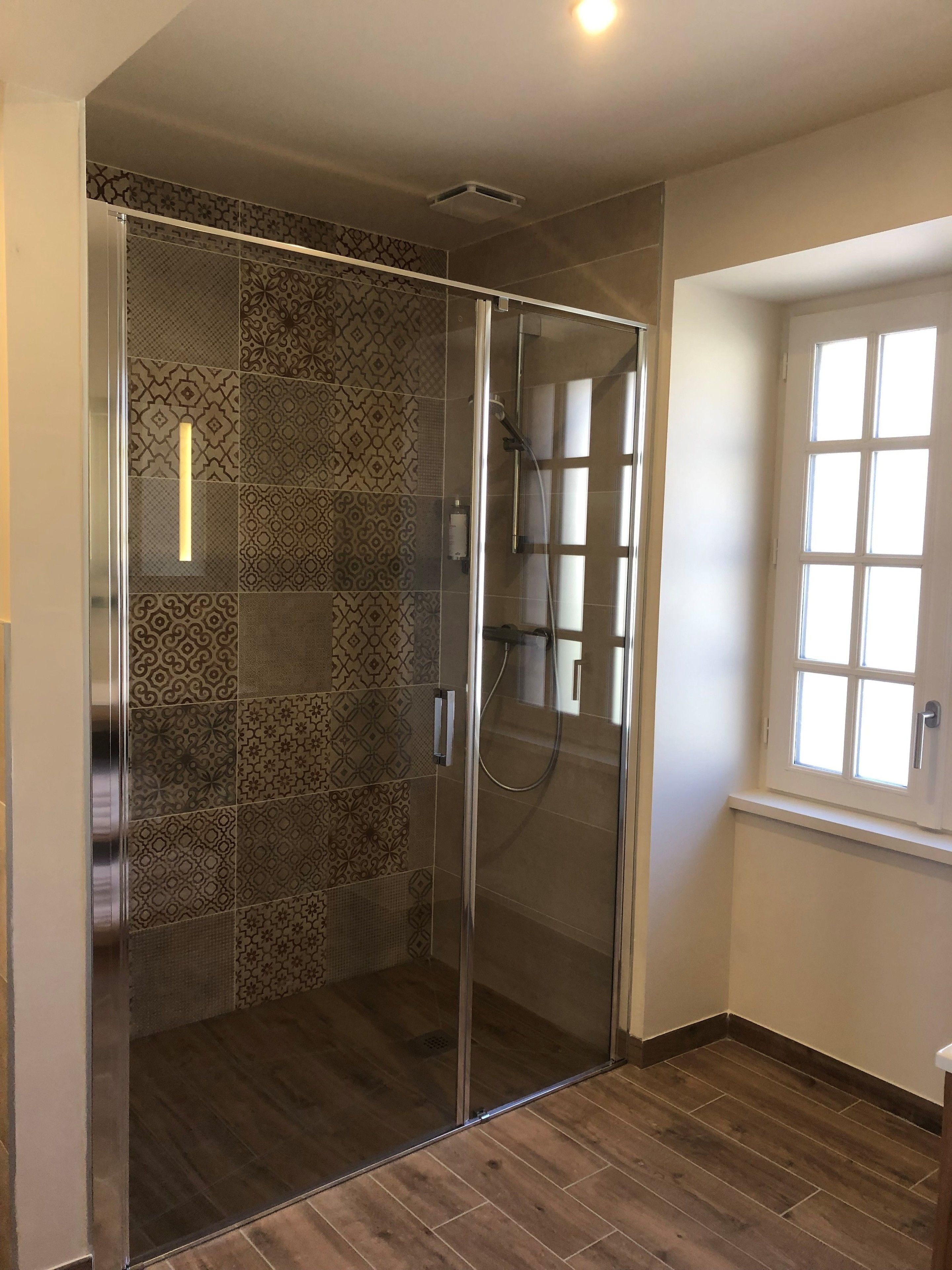 Une douche à l’italienne avec des carreaux à motifs et une porte en verre, une fenêtre avec des cadres blancs et un plancher en bois.
