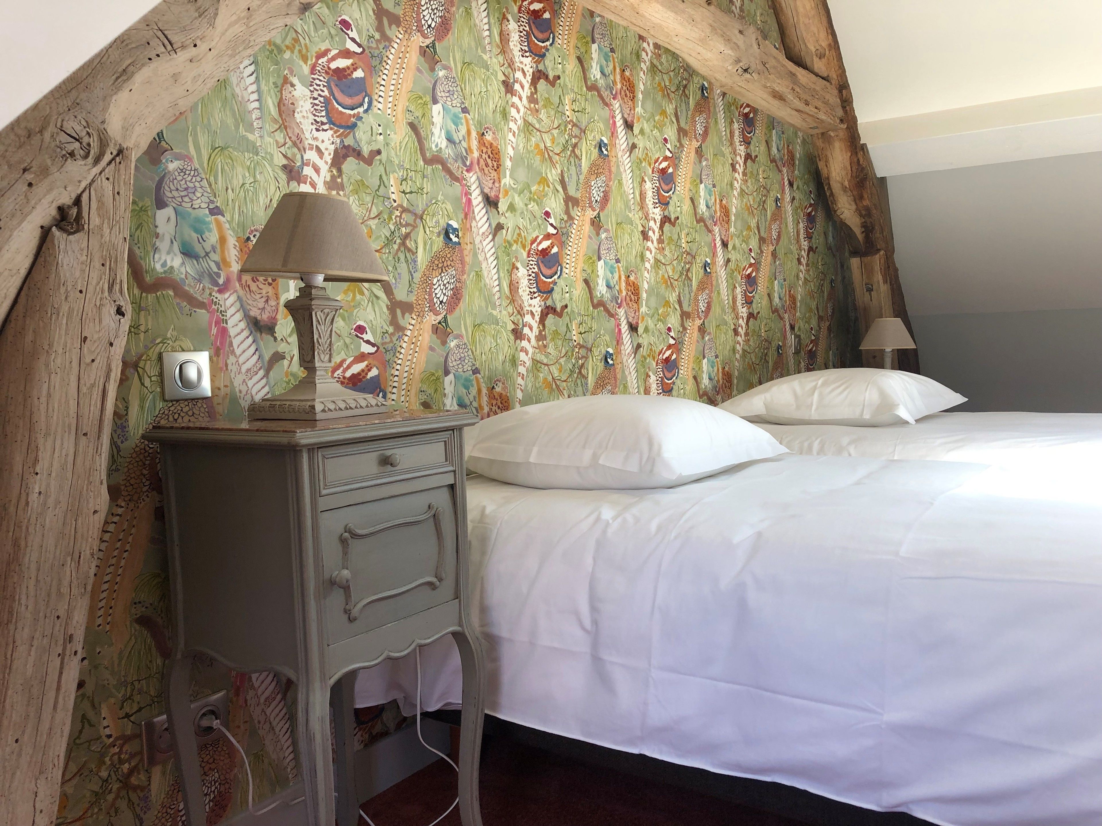 Un dormitorio con cama, dos almohadas, una mesita de noche con lámpara y una pared con diseño de pájaros coloridos.