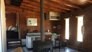 A rustic cabin interior with a central fireplace, a kitchen area, and a seating area with a sofa and chairs. - Boschetto Suites De Bosques (Athos Pampa)