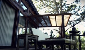An outdoor seating area with a wooden table and chairs, a covered patio, and a view of trees and a building.