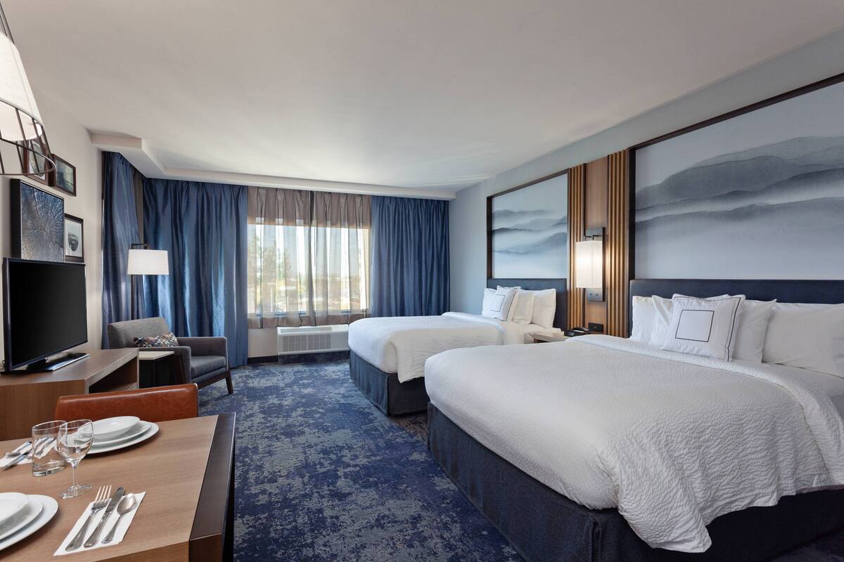 Une chambre d’hôtel avec deux lits, un bureau avec une chaise, une télévision et une grande fenêtre avec des rideaux.