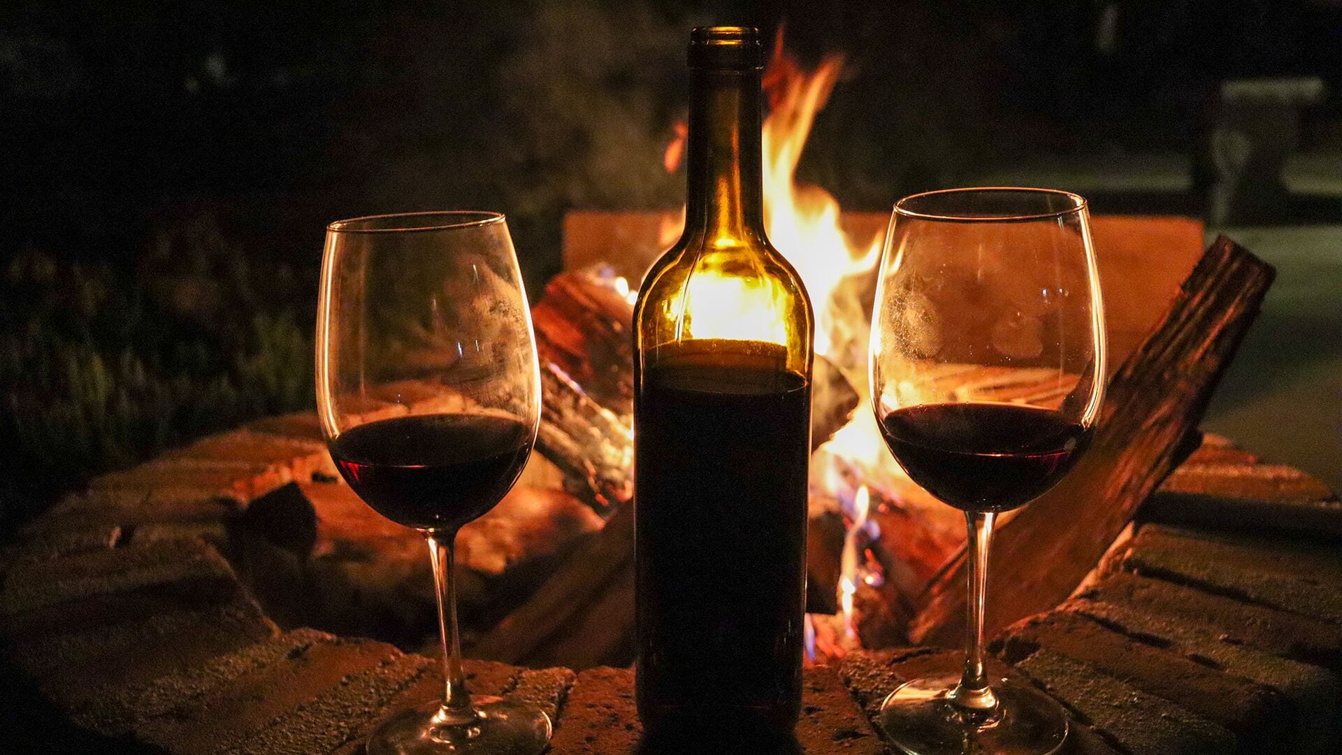 Two glasses of red wine, a bottle, and a fire in the background.