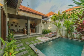 A swimming pool with a deck, surrounded by greenery and a house with a tiled roof. - Villa Dion (Canggu)