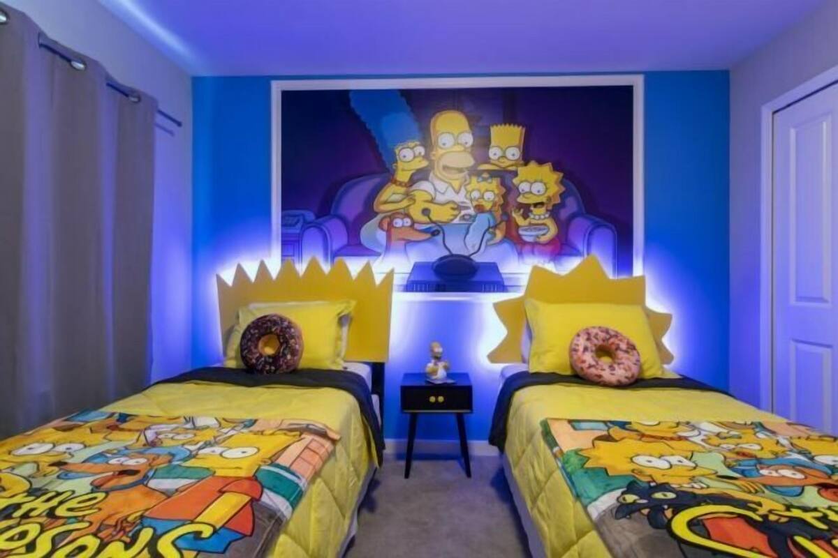 Una habitación con dos camas, un mural de Los Simpson y una mesita con una lámpara.
