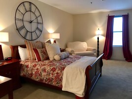 Un dormitorio con un gran reloj de pared, una cama con ropa de cama floral, dos lámparas de noche, una silla y una ventana con cortinas.