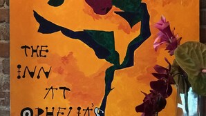 Pintura de uma flor estilizada com o texto 'THE INN AT OPHELIA'S' em um fundo de parede de tijolos.