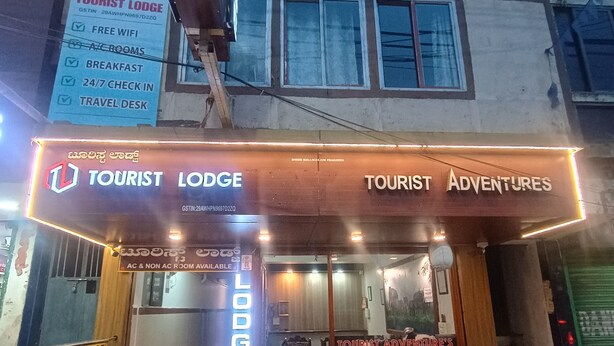 A building with a sign for 'Tourist Lodge' and 'Tourist Adventures' with amenities like free WiFi, A/C rooms, breakfast, 24/7 check-in, and a travel desk. - Tourist Lodge (Haliyal)