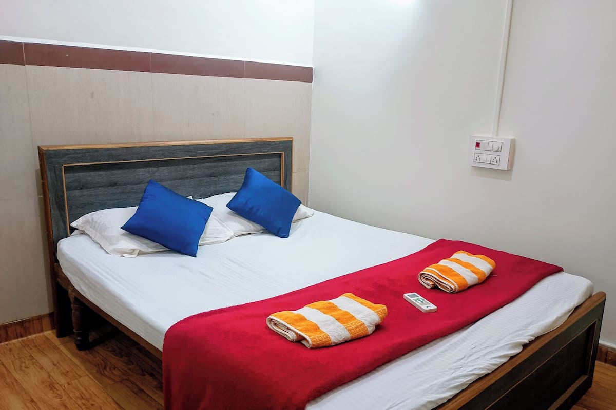 A neatly made bed with a wooden headboard, white bedding, two blue pillows, a red blanket, and two rolled towels.