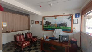A reception area with a checkered floor, wooden chairs, a desk with a computer, and a wall-mounted painting of wildlife. - Tourist Lodge (Haliyal)