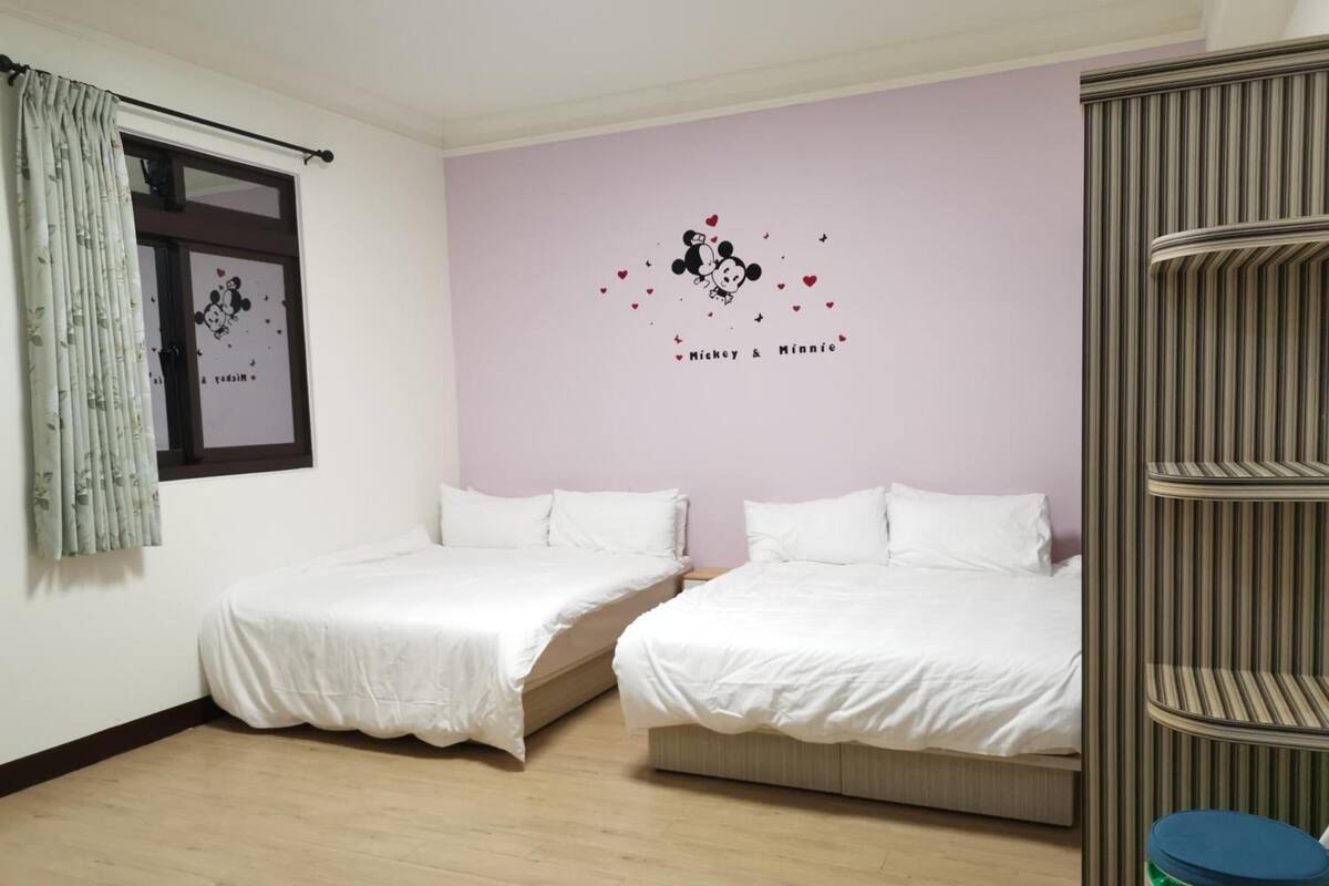 A room with two beds, a window with a curtain, a wall with a Minnie Mouse decal, and a striped cabinet.