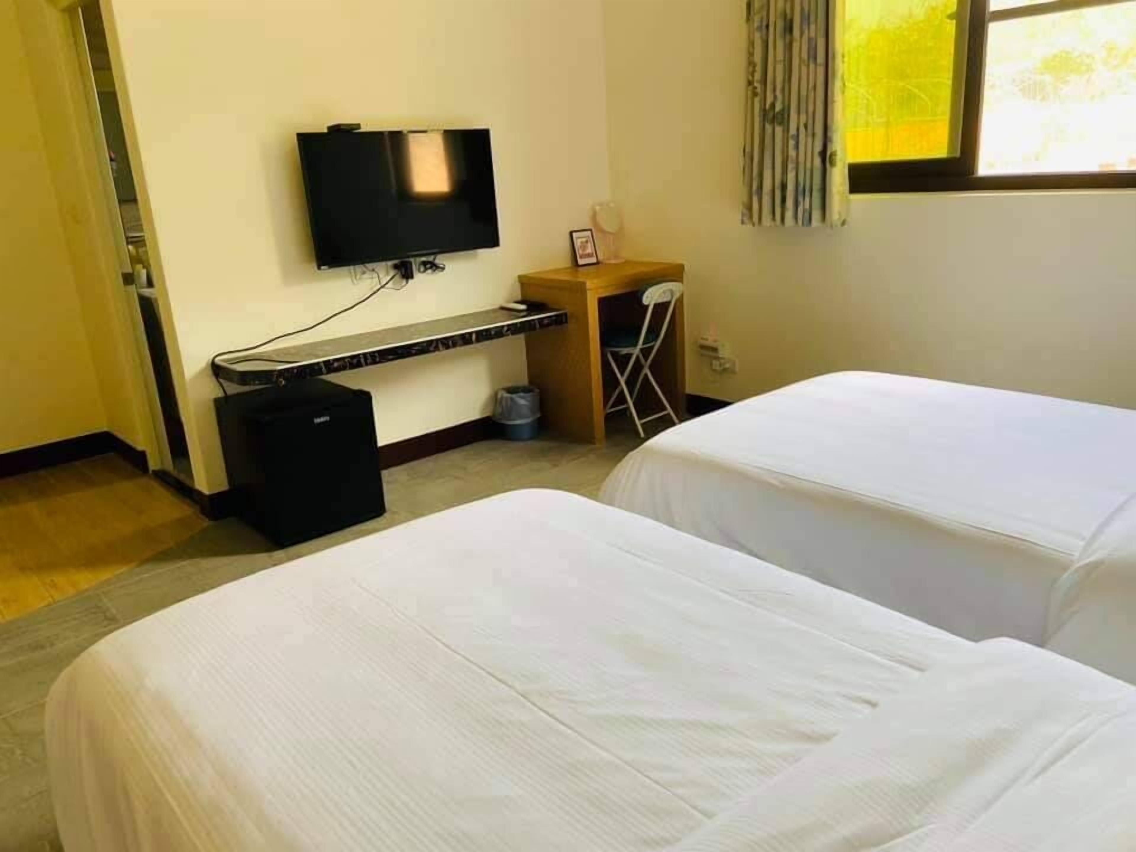 A hotel room with two beds, a desk, a television, and a window with curtains.