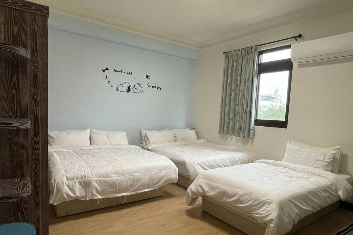 A hotel room with two single beds, a wooden wardrobe, a blue wall with a 'Good night' and 'Snoopy' decal, and a window with curtains.