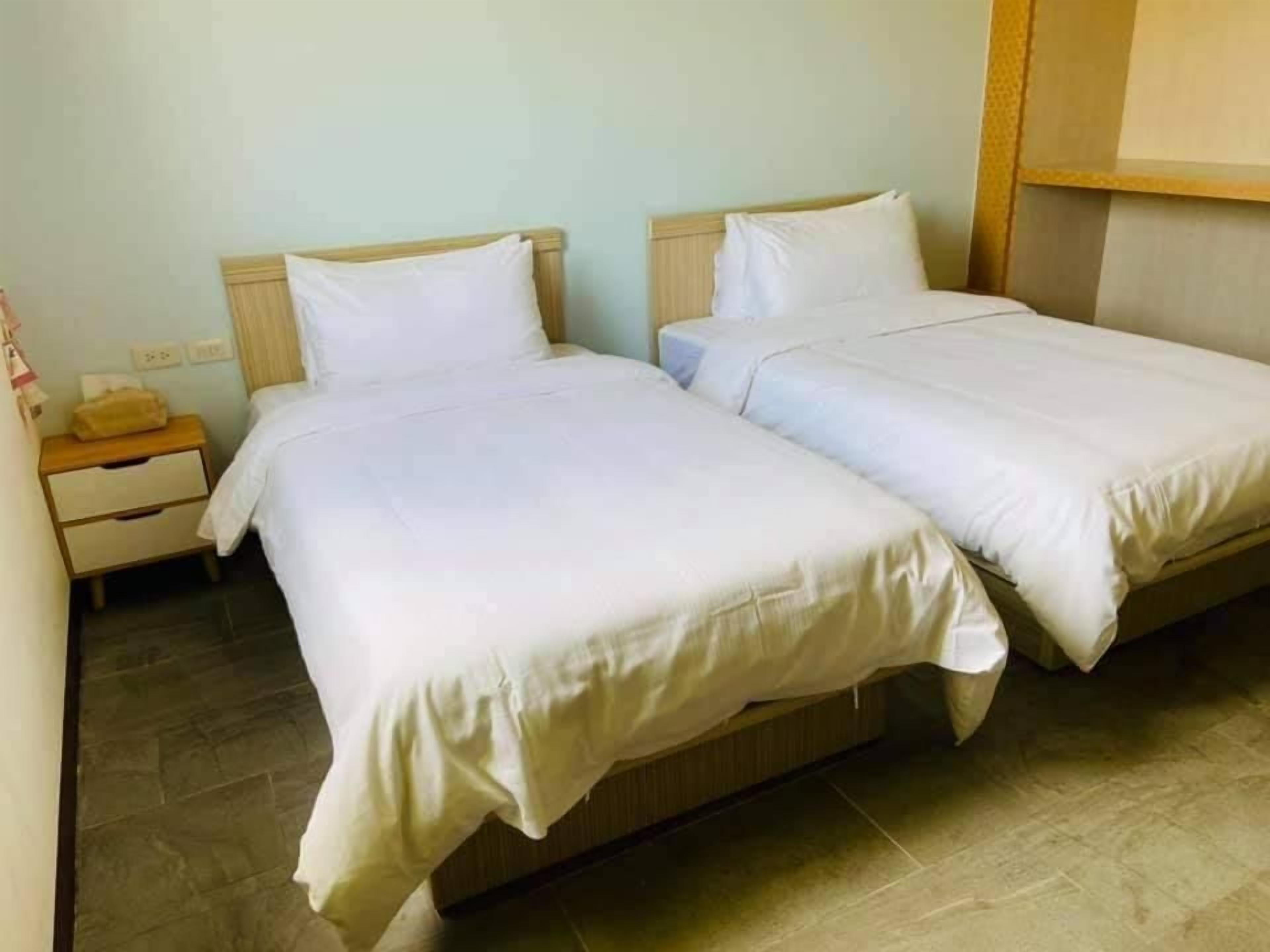 A room with two single beds, a wooden headboard, a nightstand with a lamp, and a tiled floor.