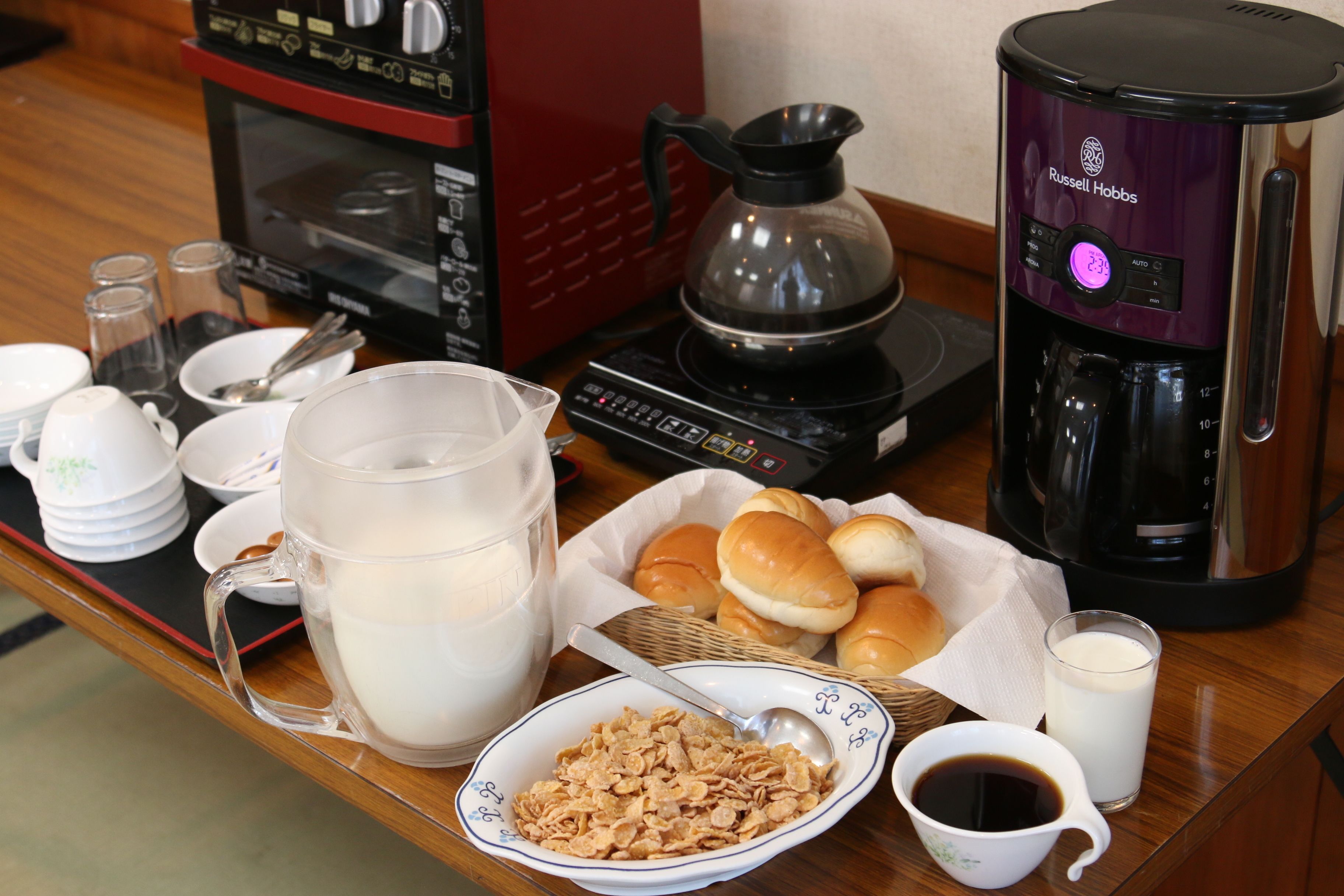 A breakfast setup with a coffee maker, milk jug, cereal bowl, and a basket of rolls.