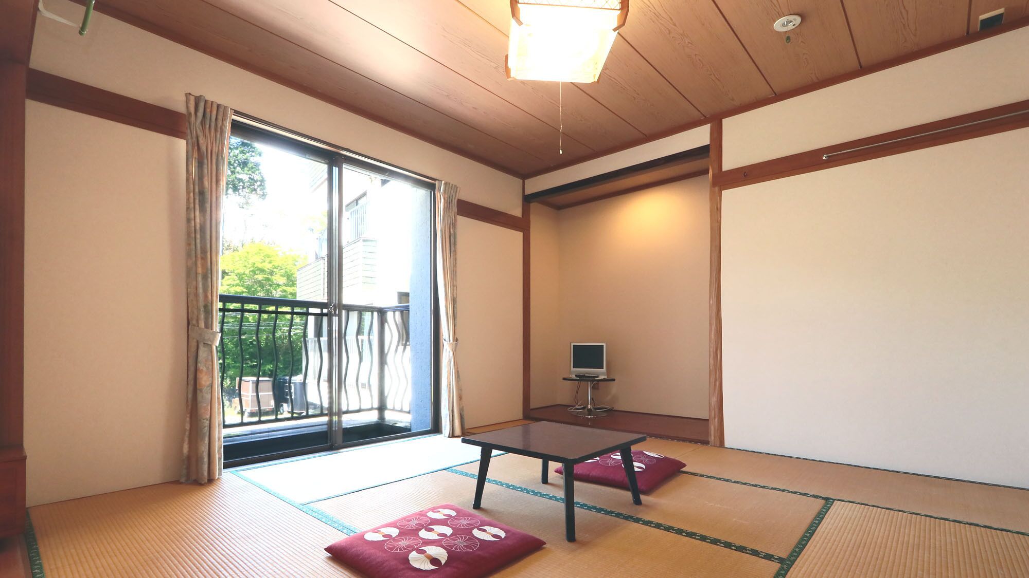 A traditional Japanese room with tatami flooring, a sliding door, a small table with a TV, and a balcony with a view of greenery.