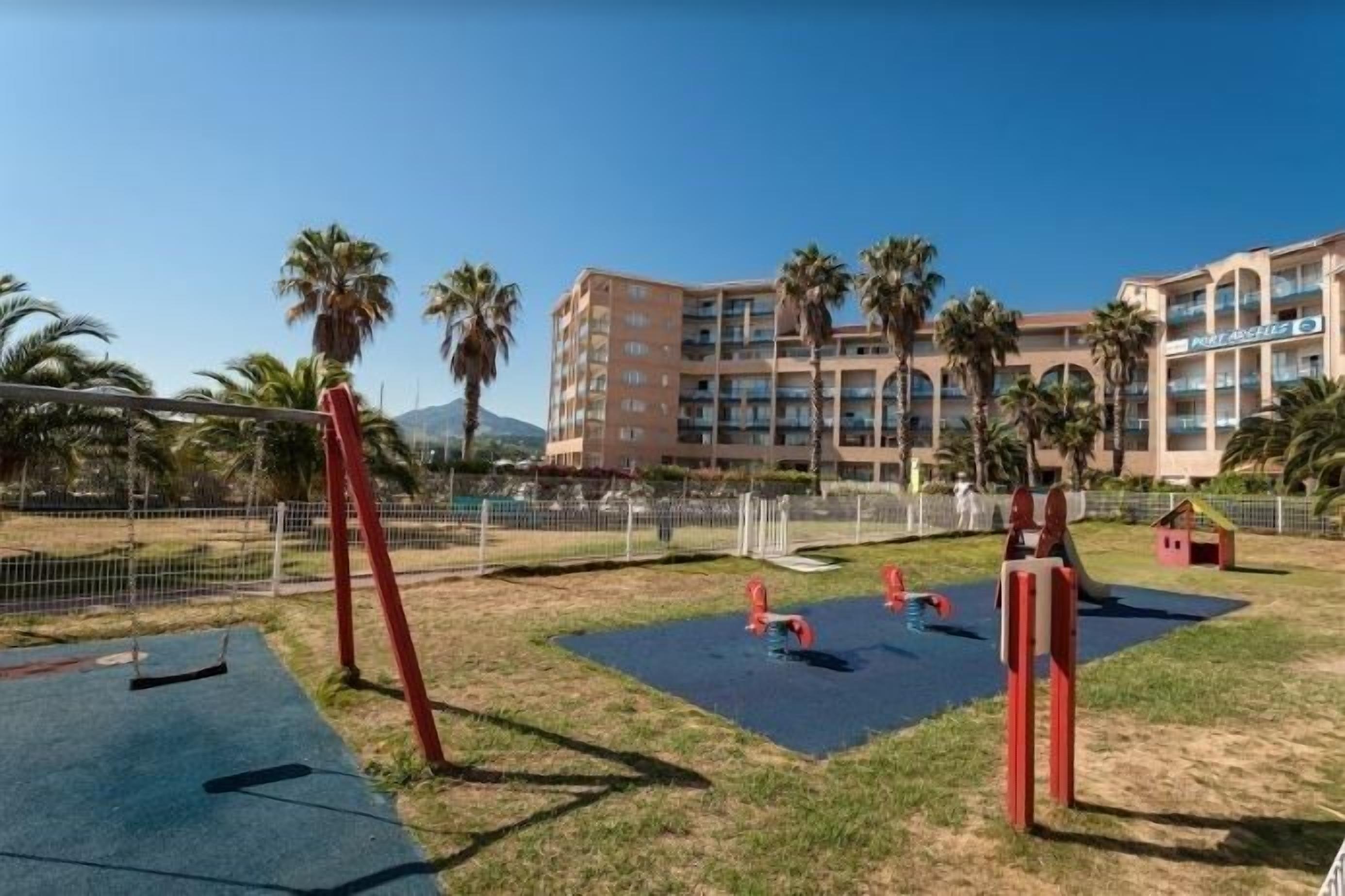 A playground with swings and slides, surrounded by a fence, with a hotel building in the background.