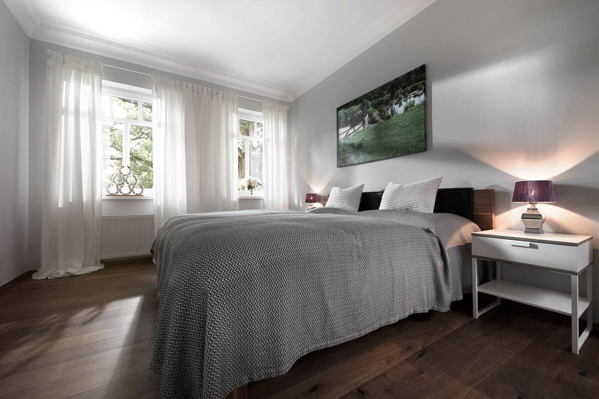 A bedroom with a large bed, white curtains, a nightstand with a lamp, and a painting on the wall.